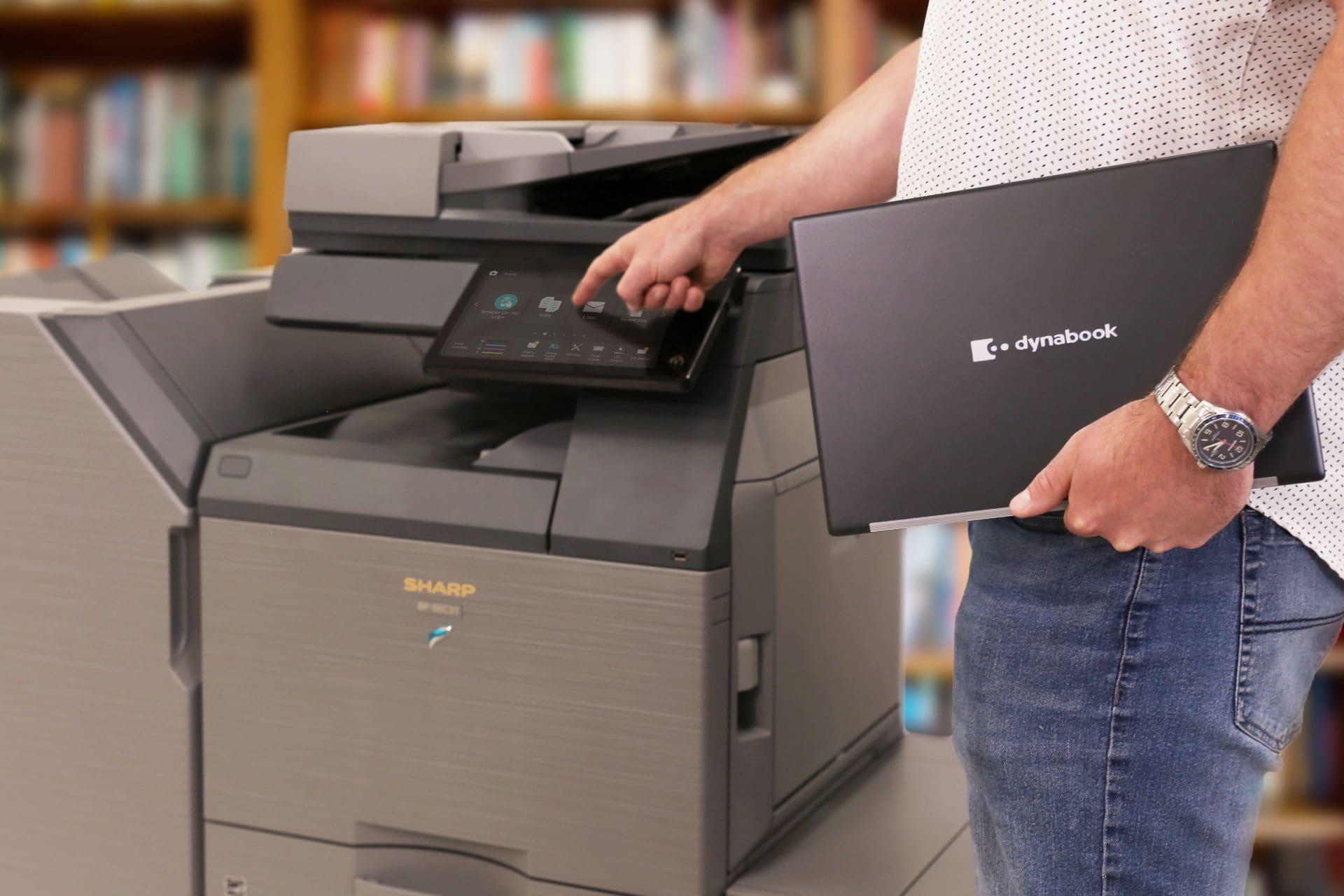 Man in a dotted shirt holds a Dynabook laptop next to a large Sharp multi-function printer in a library setting, with his watch visible on his wrist.
